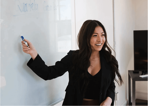 A woman in black jacket writing on white board.